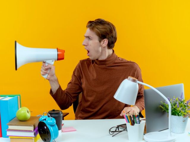 looking-side-young-student-boy-sitting-desk-with-school-tools-speaks-loudspeaker_141793-80508