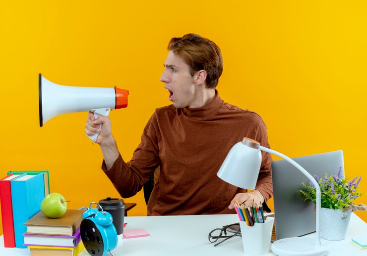 looking-side-young-student-boy-sitting-desk-with-school-tools-speaks-loudspeaker_141793-80508